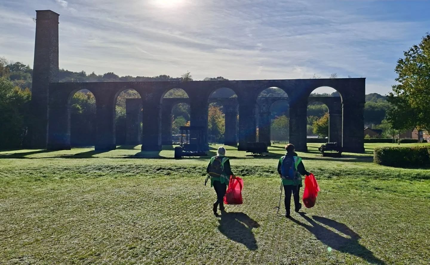 Two volunteers with litter pickers and red bags walking towards historic stone arches on a sunny morning