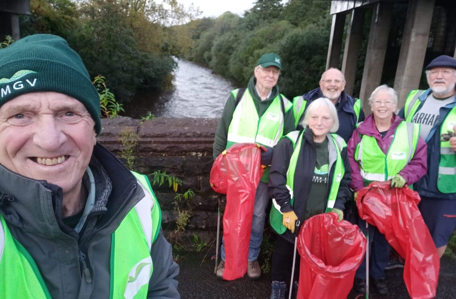Smiling volunteers in hi-vis with red bags on a bridge over the river, ready for a litter pick